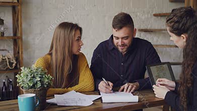 Attractive broker is selling house to young couple, people are signing documents and shaking hands, realtor is giving