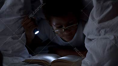 Child reading books at night under blanket, lighting himself with flashlight
