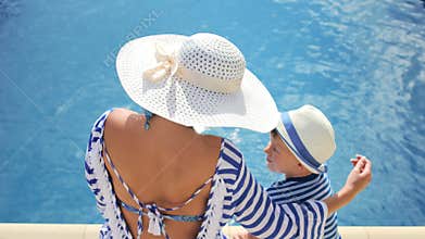Woman in white hat with bow and little boy sitting at edge of swimming pool together