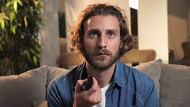 Sad young man holds a remote control in his hands and switches channels on TV.