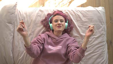 Happy teen girl with pink hair wearing headphones lying in bed listening music.
