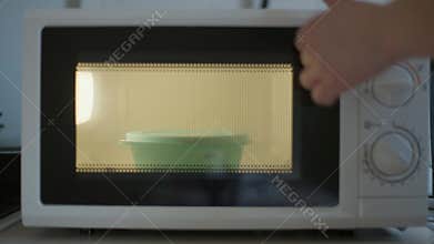 White microwave oven on table with female Caucasian hand putting food container with remains inside closing door turning