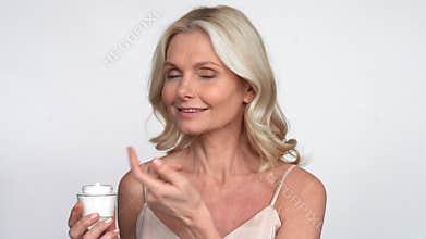 Smiling 50s middle aged woman putting facial cream on face looking at camera.