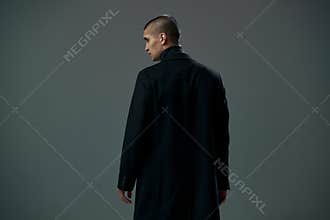 Brave young man dressed in black coat detective or criminal on gray studio background, back view