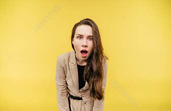 Portrait of shocked girl in a beige jacket on a yellow background, looking at the camera with a funny face