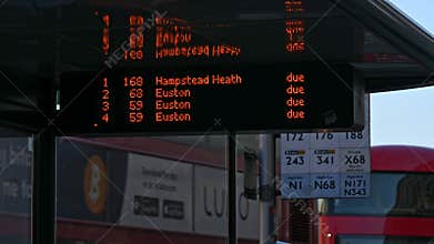 A close up of a digital sign in a bus stop saying which buses are due as Red London Double Decker Buses pass in the background