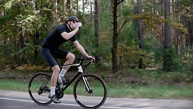 A male cyclist drinks water riding a road bike in a pine forest, wearing helmet
