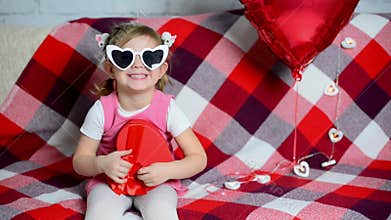 Little beautiful girl is sitting on sofa with glasses and smiling sweetly. Red heart-shaped gift box and balloon. Festive atmosphe