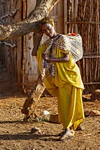 A girl carrying a jerry can of water on her back rests her head on a tree to rest.
