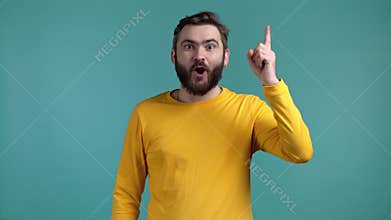 Smiling ingenious guy showing eureka gesture. Portrait of man having idea moment pointing finger up on blue studio
