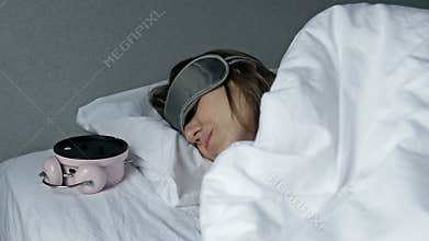 Woman hates waking up early in the morning. Sleepy woman looking at alarm clock and trying to hide under the pillow