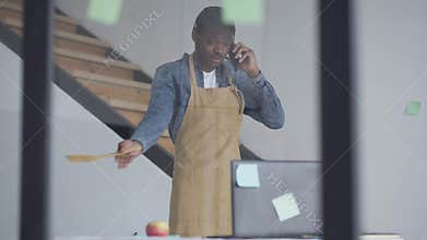 Busy man in apron talking on the phone gesturing with cooking shovel. Portrait of confident African American employee