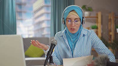 Portrait young muslim woman records a podcast while sitting in front of a microphone in her living room