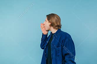 Young man in a blue shirt with a long hair pelvis hides to the side covering his palm on a blue front. Copy space