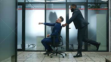 Two funny businessmen riding office chair while having fun in lobby of modern business center. Business people celebrate