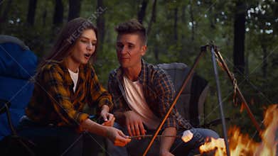 Camping and picnic concept. Young affectionate man and woman frying marshmallow on fire, enjoying evening in forest