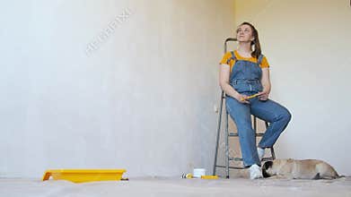 Happy woman with brush and ladder looks dreamily in a room with an unpainted wall with copy space. Independent single