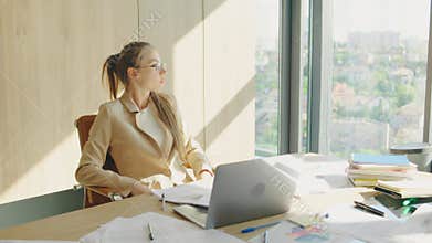 A focused businesswoman is working at a modern office with a scenic city view. She is busy with paperwork, using a