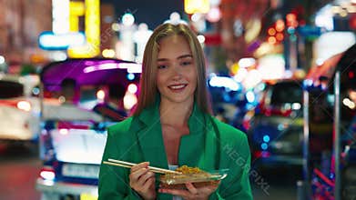 Portrait of pretty woman with noodle box looking at camera standing in night street market.