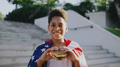 Under the sunny sky, a cheerful, patriotic boy joyfully eats a burger wrapped in the American flag outdoors, celebrating