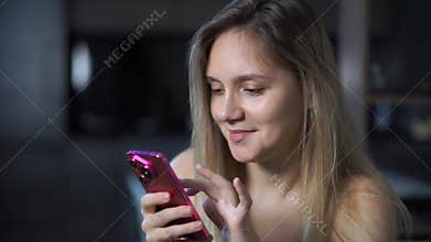 young woman with long hair is happily engaging with her pink smartphone. She is wearing a simple white t-shirt. A girl