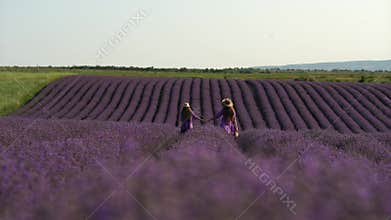 Mother and daughter walk through a lavender field dressed in purple dresses, long hair flowing and wearing hats. The