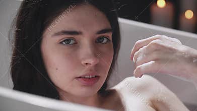 A woman is relaxing in a bathtub with a candle glowing in the background. Blow foam from hand and look at camera