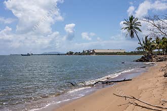 A tropical beach with palm tree, blue sky and clouds and a sugar terminal with jetty