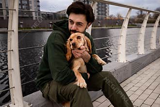 A young brunette guy poses with his dog on the background of the river. A man sits on the street and hugs his cocker