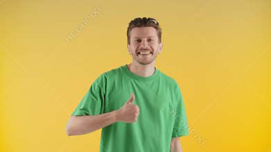 Smiling man in green t-shirt showing thumbs up. A man stands against a yellow wall and smiles at the camera.