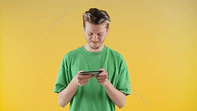 A cheerful young guy in a green T-shirt stands near a yellow wall and plays on the phone. A young man is having a good