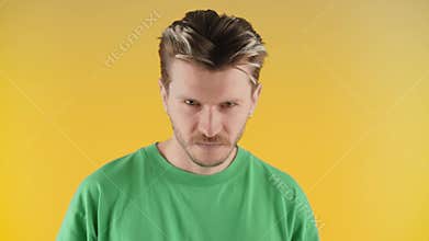 A frowning man with stubble is looking at the camera while standing on a yellow background. A young man in a green T