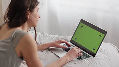 Rear view of a young woman typing on a green screen laptop. A young brunette woman works remotely on a laptop while