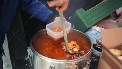 Serving soup in a in a plastic container, A person in a blue jacket ladles soup into a container from a large pot at a