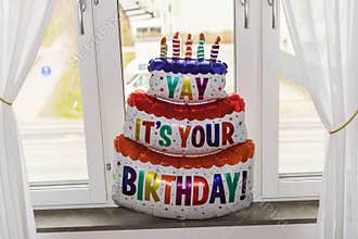 Close-up view of a vibrant birthday balloon shaped like a cake with candles and a festive greeting by a window.