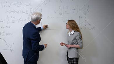 An elderly professor explains a subject to a student at a white board.