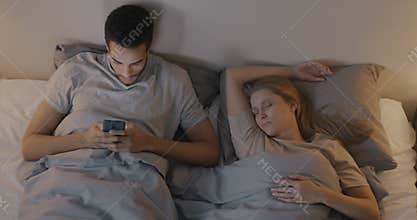 African American man using smartphone texting and looking at sleeping wife Caucasian woman in bedroom in evening