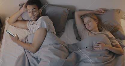 African American guy using mobile app messaging with smartphone and looking at sleeping wife lying in bed in evening