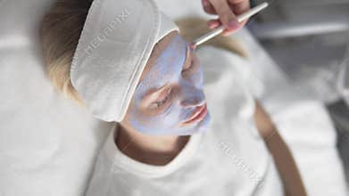 The cosmetologist applies a blue moisturizing mask to the face of a female client.