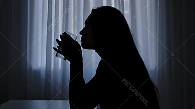 Silhouette of woman sitting at table in front of window, shocked has hand tremor, drinks glass of water, mental