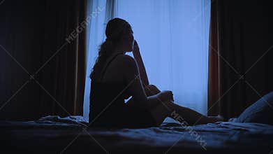 black silhouette upset unhappy woman sitting on bed dark room, cry wipes away tears with hand mood depression sadness
