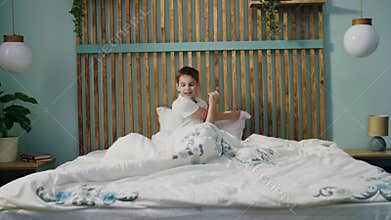 A little boy wakes up, stretches, does morning exercises with crosses arms, rubs eyes with hands and yawns, sitting on