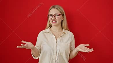 Baffled young blonde woman wearing glasses standing over a red background, her clueless, confused expression oozing doubt