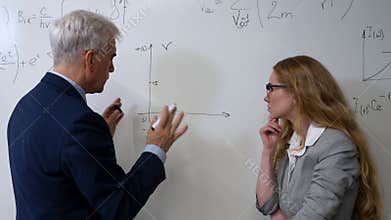 An elderly professor explains a subject to a student at a white board.