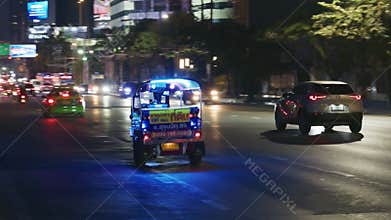 Traditional Tuk Tuk taxi driving at night on a bustling Bangkok street with modern city traffic and buildings in a