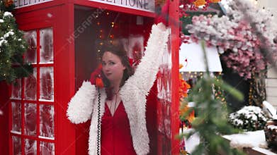 A Christmas concept, Happy woman in a fur coat posing in a red telephone booth on snowing day.