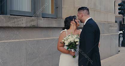 A beautiful plus-size bride stands on a city street smiling with a wedding bouquet, as the groom approaches her and they gently
