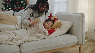 Mother decorates a Christmas tree in a cozy apartment while her daughter sleeps peacefully on the sofa. She gently covers her with