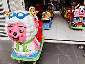 A young child wearing an orange outfit with a beige hijab is seated on a coin-operated ride shaped like a colorful cartoon sheep.