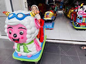 A young child wearing an orange outfit with a beige hijab is seated on a coin-operated ride shaped like a colorful cartoon sheep.